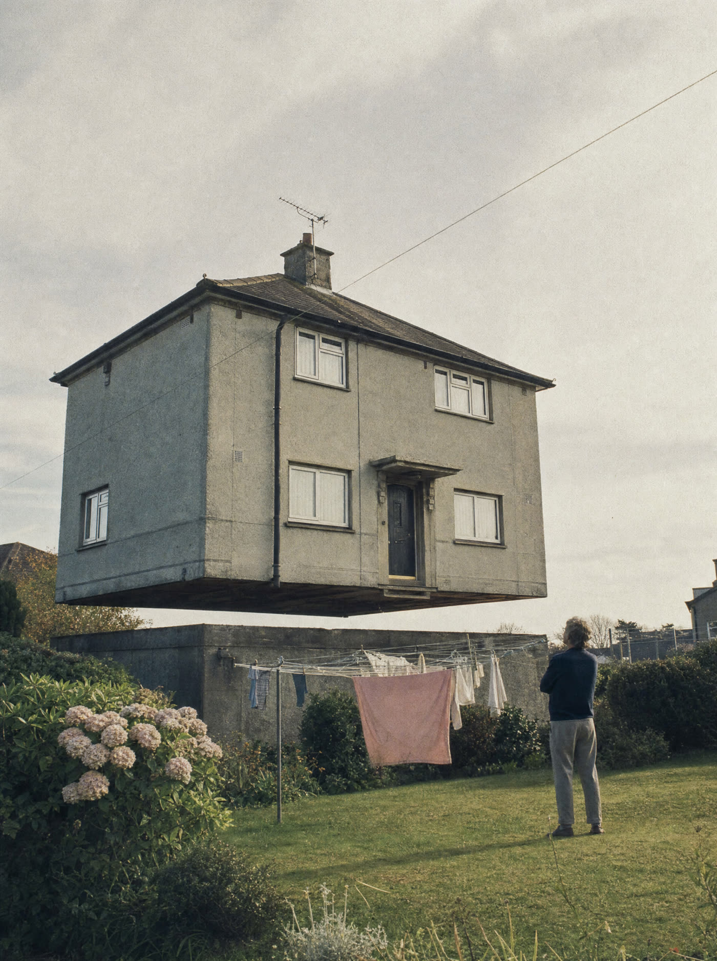 A suburban house floating a metre above its own foundations, a resident on the lawn looking at it