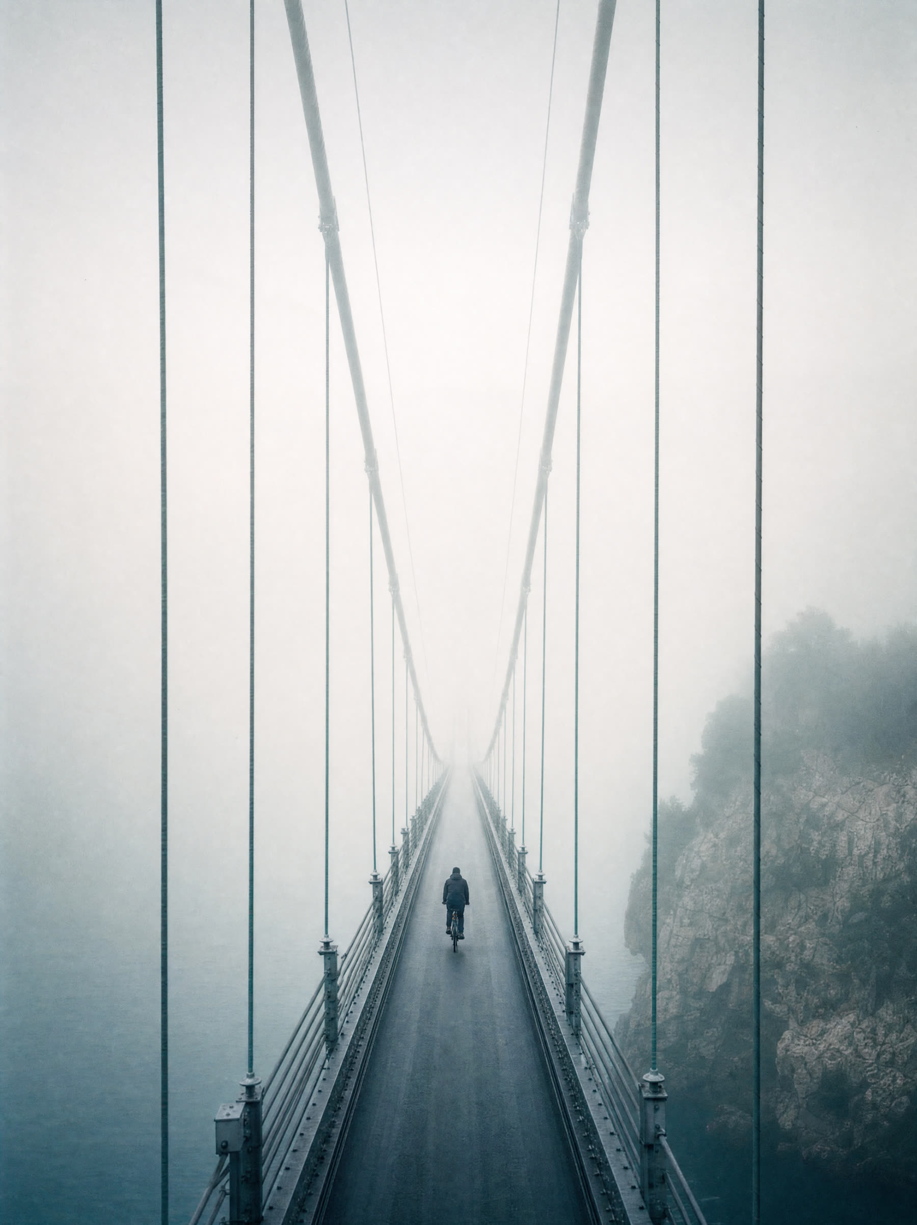 A suspension bridge vanishing into thick sea fog, one cyclist alone on the deck